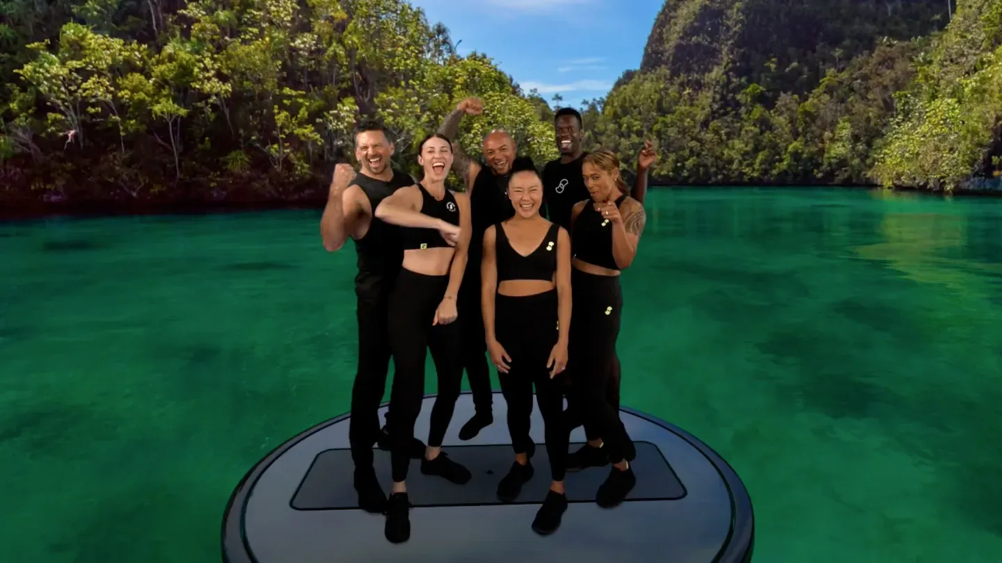 The six Supernatural coaches cheering together on a round workout platform floating on a green tropical lagoon ringed by forested cliffs